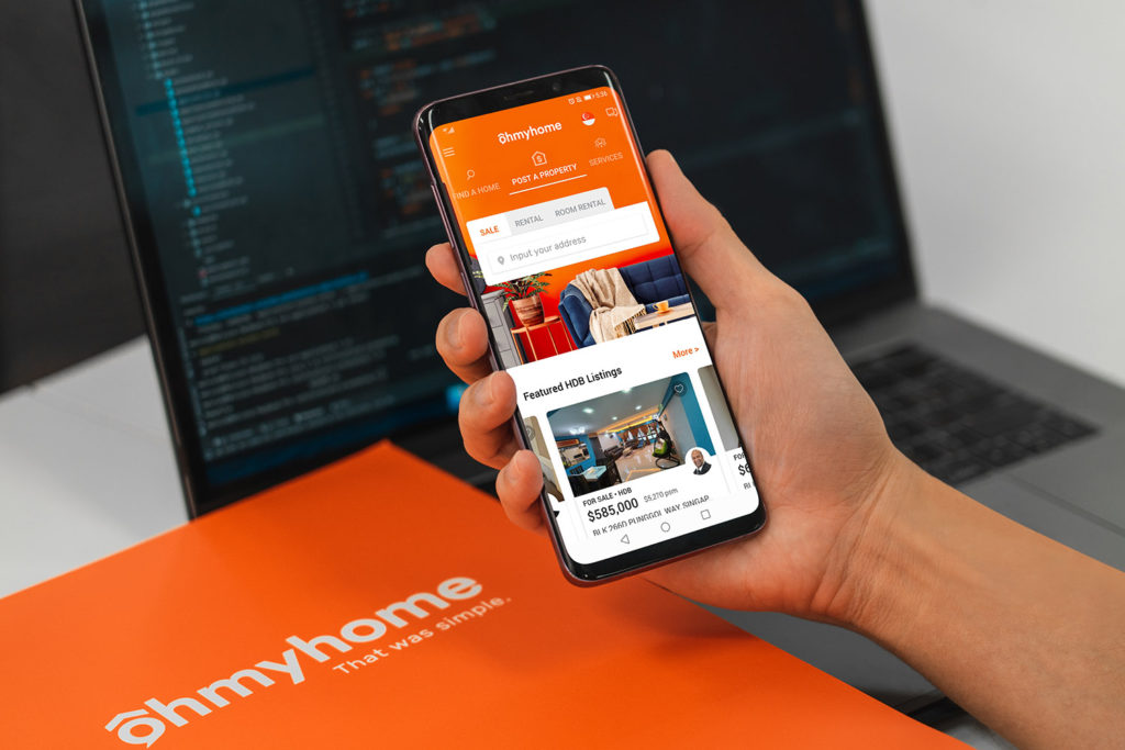 A hand holding a phone showing the Ohmyhome app