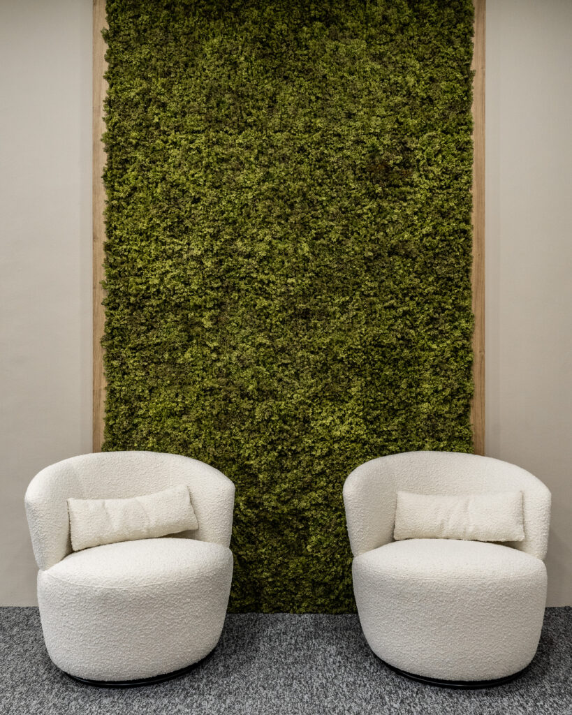 A stylish office lounge area featuring two plush white chairs positioned in front of a vertical green moss wall. The textured greenery adds a natural and serene element to the space, complementing the cozy seating and creating a relaxing ambiance for informal meetings or quiet breaks.