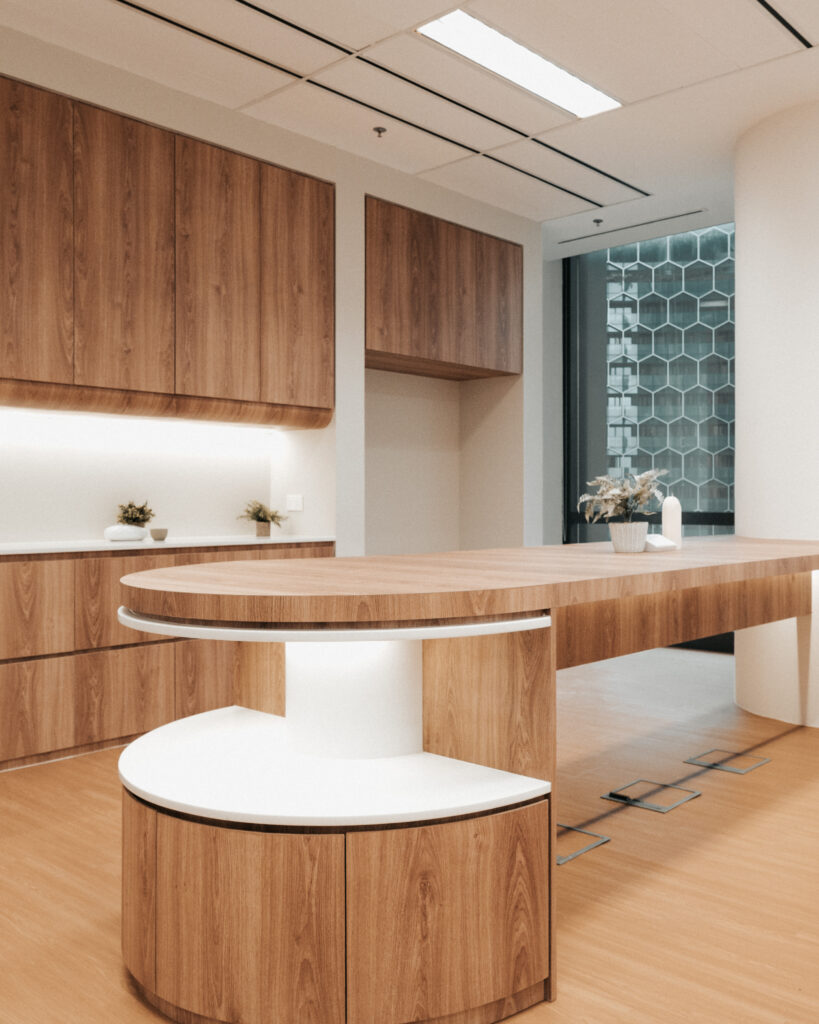 A modern office kitchen area with elegant wood cabinetry and a stylish central island. The space features a mix of closed storage and open shelves, decorated with small plants and minimalistic vases. Natural light from large windows enhances the warm and inviting atmosphere, complemented by the clean lines and contemporary design.