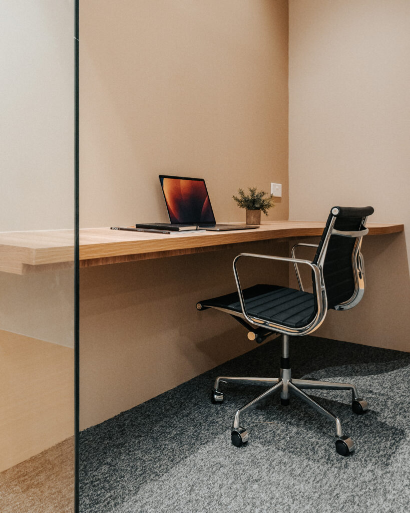 A cozy and modern private workspace featuring a minimalist wooden desk and an ergonomic chair. The desk holds a sleek laptop and a small potted plant, creating a clean and focused environment. The area is well-lit and enclosed by a glass partition, providing both privacy and an open feel.