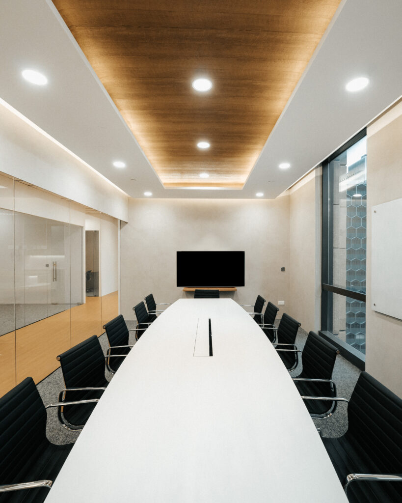 A modern conference room featuring a long white table surrounded by sleek black chairs. The room is illuminated by recessed lighting and has a wooden accent on the ceiling, adding warmth to the minimalist design. A large screen is mounted on the wall at the far end, perfect for presentations and meetings, with glass walls allowing natural light to flow in.