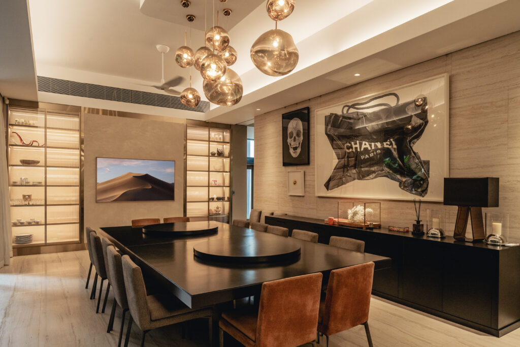 A modern dining room features a large, dark wood table surrounded by brown leather chairs. The room is illuminated by a cluster of spherical pendant lights and a wall-mounted television. Artwork adds a touch of personality to the space, showcasing a Chanel bag and a skull.