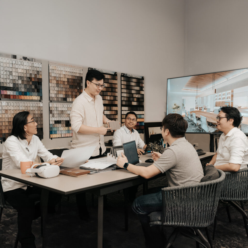 The image depicts a meeting at a renovation firm in Singapore, featuring a collaborative team of interior designers, architects, and project managers. One person stands at the head of the table, leading the discussion, while others are seated, actively engaging with the presentation and materials around them. The backdrop includes various sample materials and a large screen displaying a modern interior design, reflecting their focus on creating well-planned, aesthetically pleasing spaces.