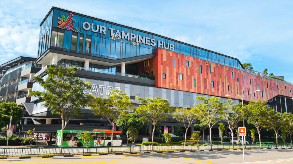 A modern, multi-story building with colorful accents and a large glass facade, identified as "Our Tampines Hub". A tree-lined street with a public bus passes in front of the building.